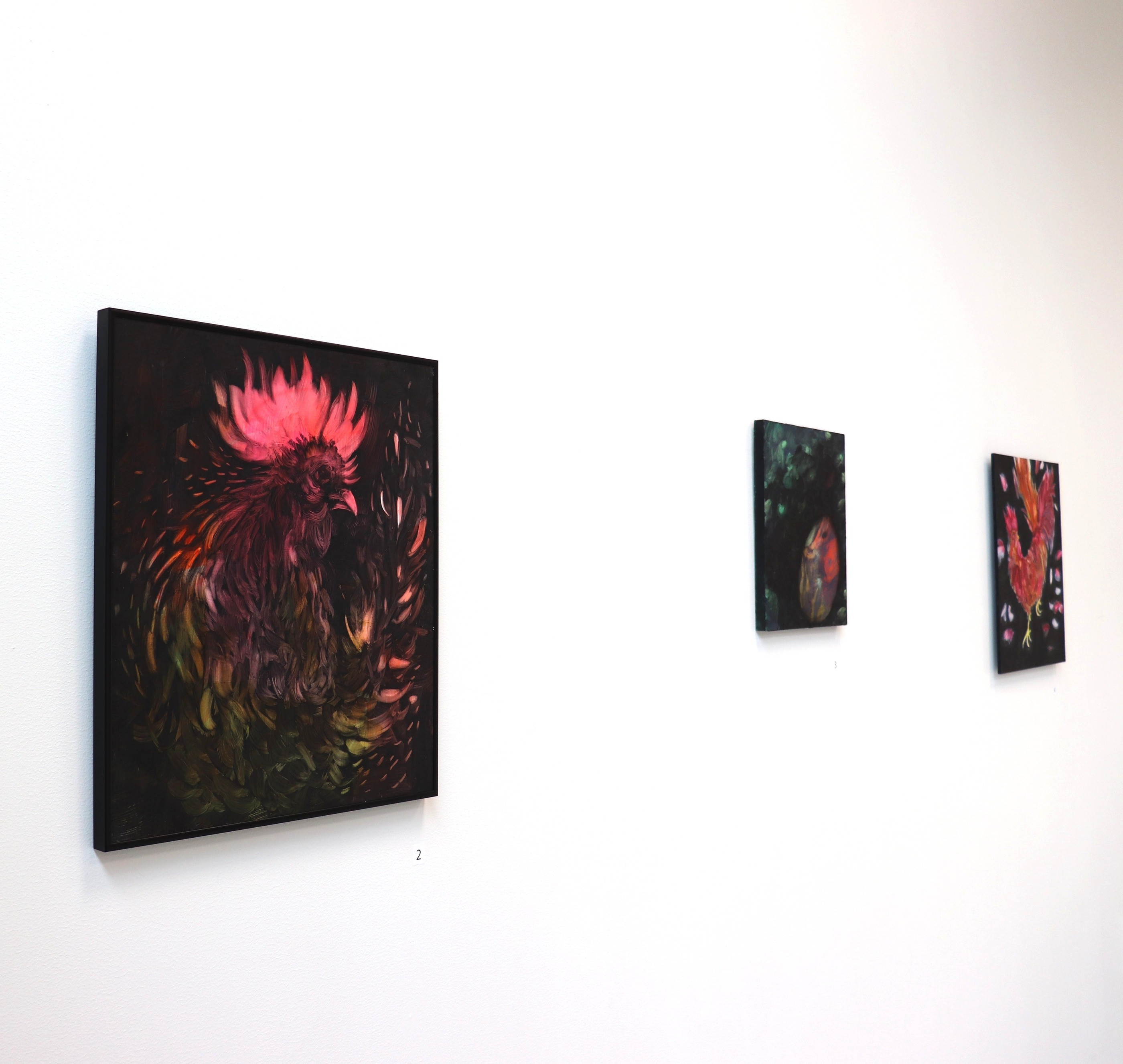 A white gallery wall with a large framed painting of a rooster crowned in pink, and two smaller dark works further along.