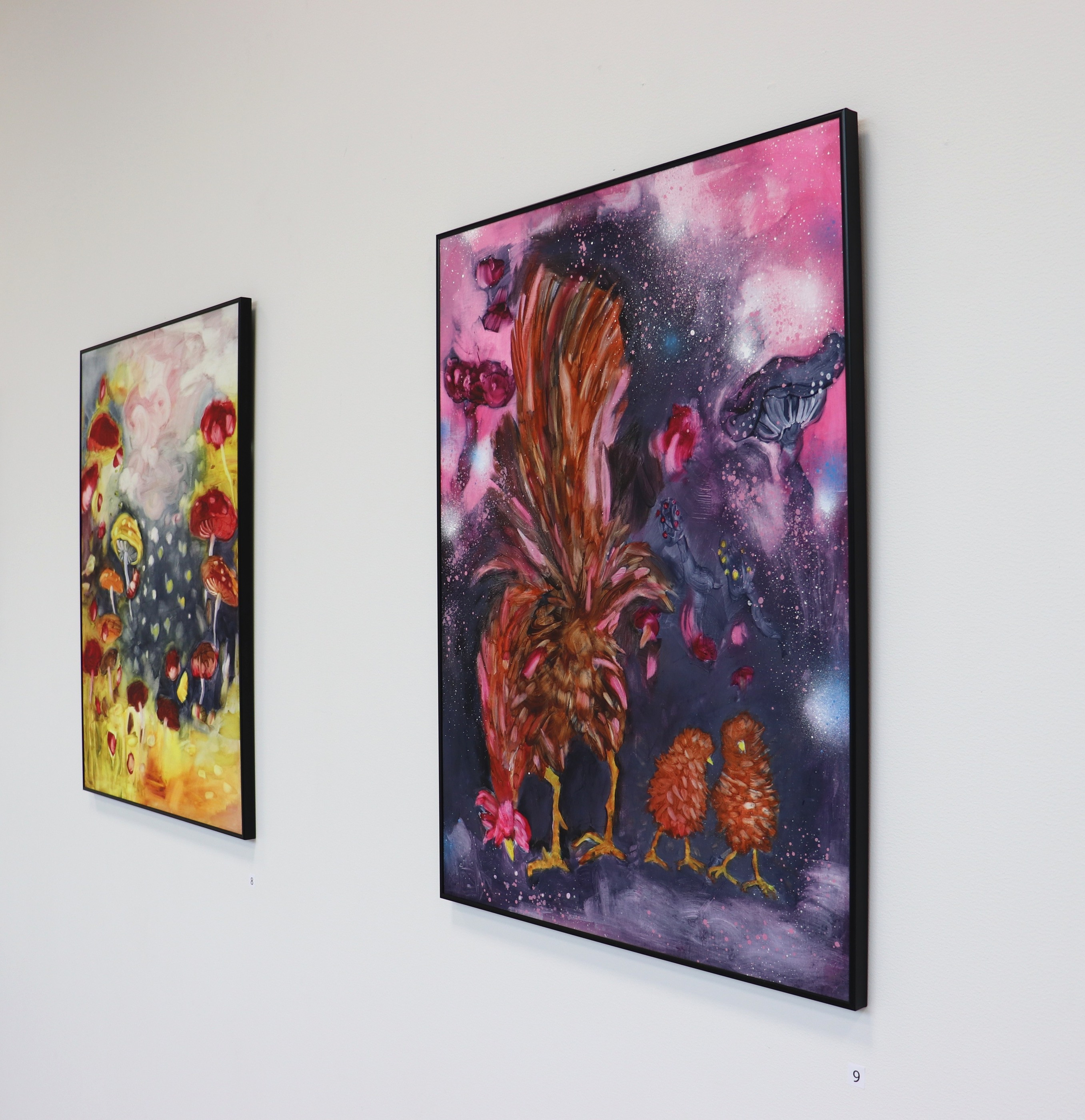 An angled gallery view of a red and yellow mushroom painting beside a larger work of a russet rooster and two chicks in pink and violet.