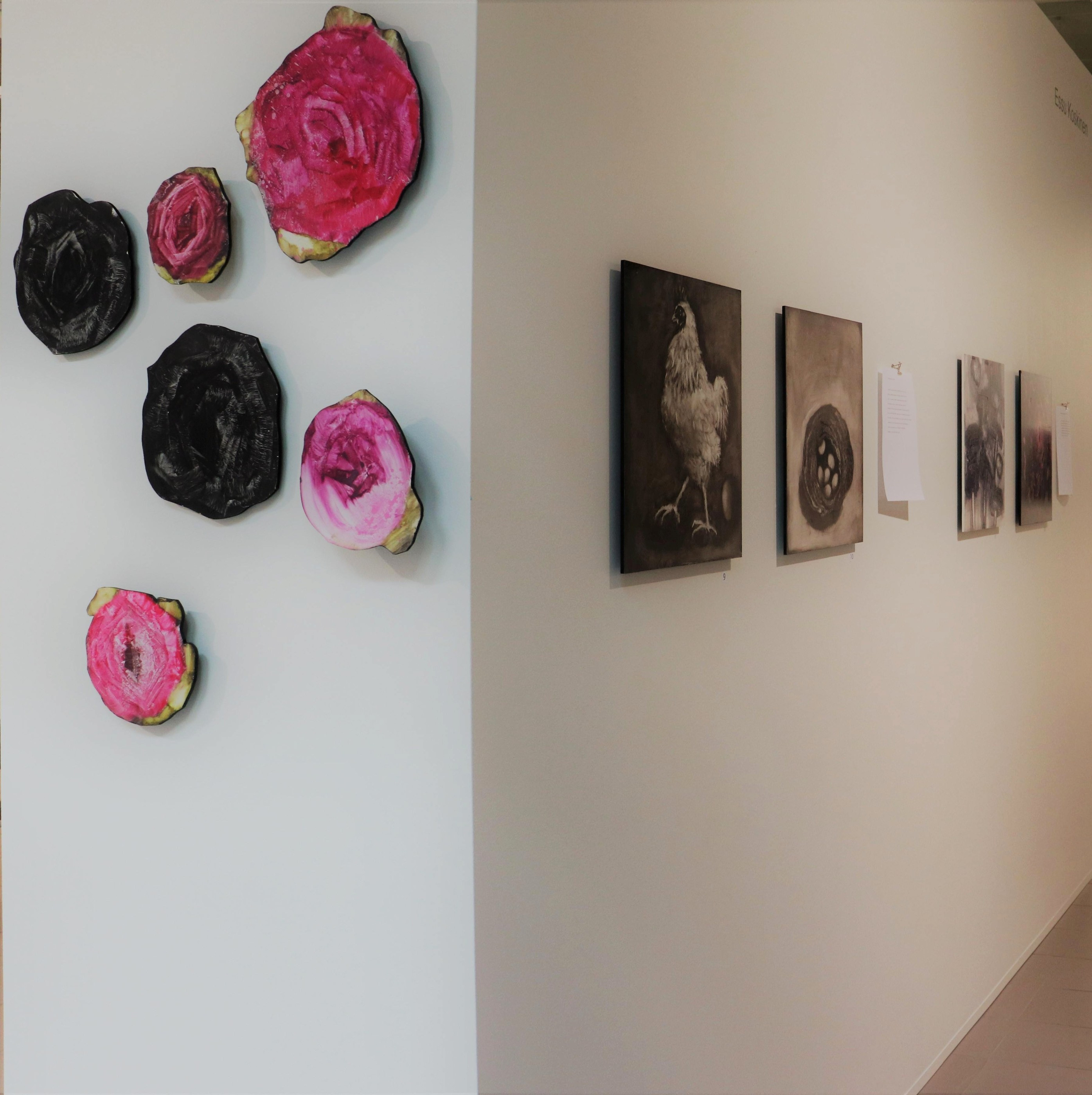 A gallery corner with sculptural pink and black flowers and a sequence of small dark works.