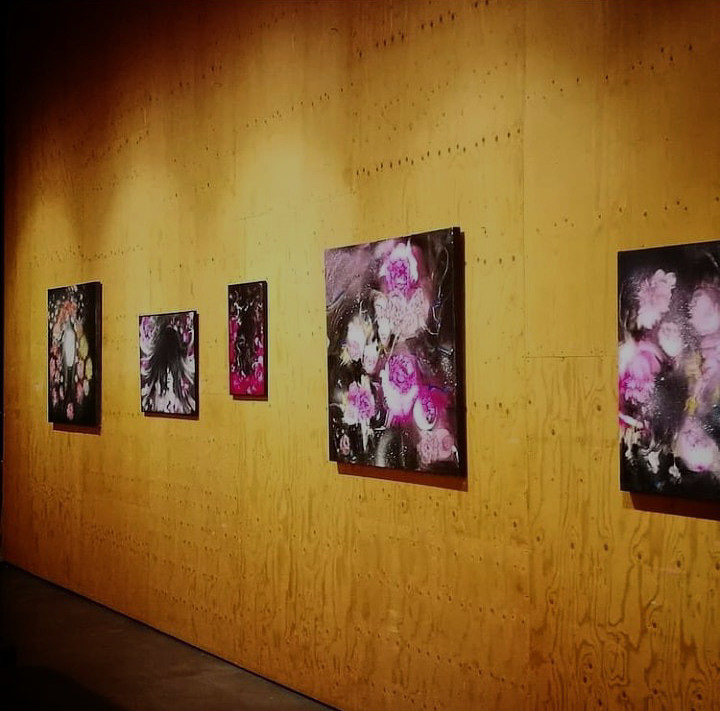A sequence of dark floral paintings installed along a warm wood-paneled gallery wall.