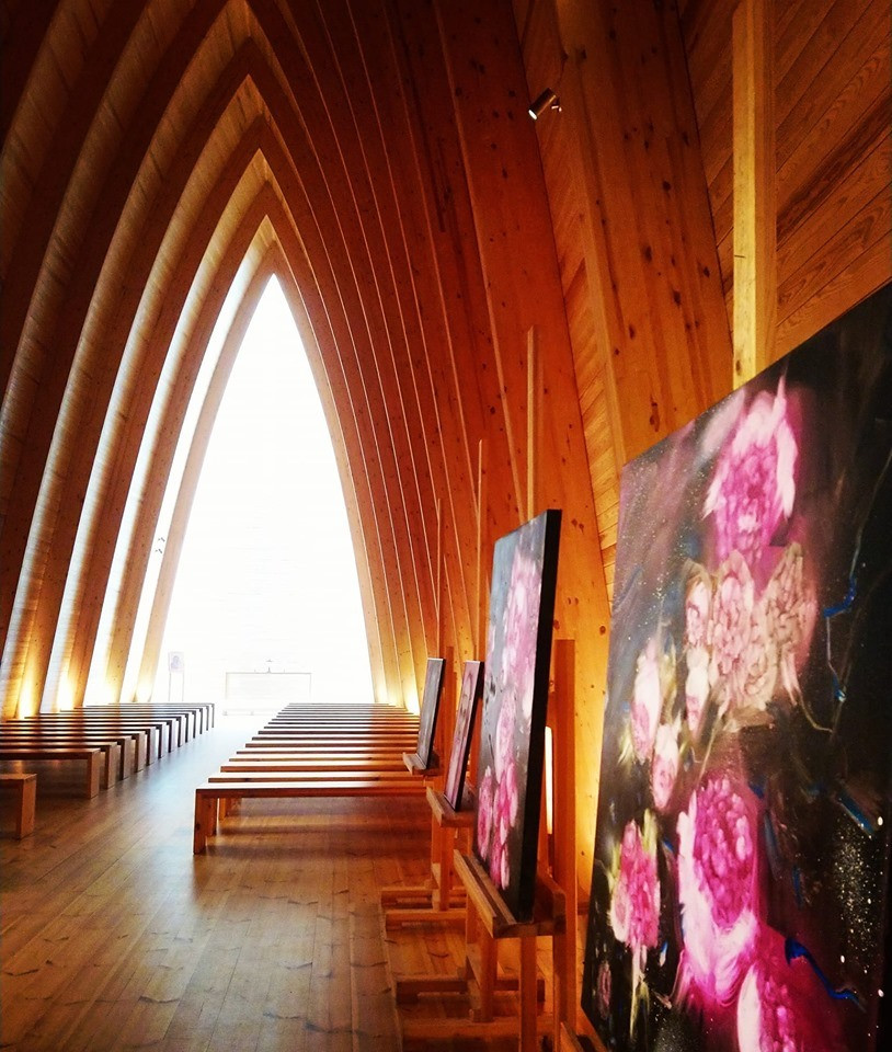 Paintings displayed inside a wooden chapel beneath a dramatic row of pointed windows.
